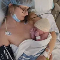 Courtney with theatre gown and cap, ECG pads, and a blood pressure cuff holding her newborn baby Adam Gordon